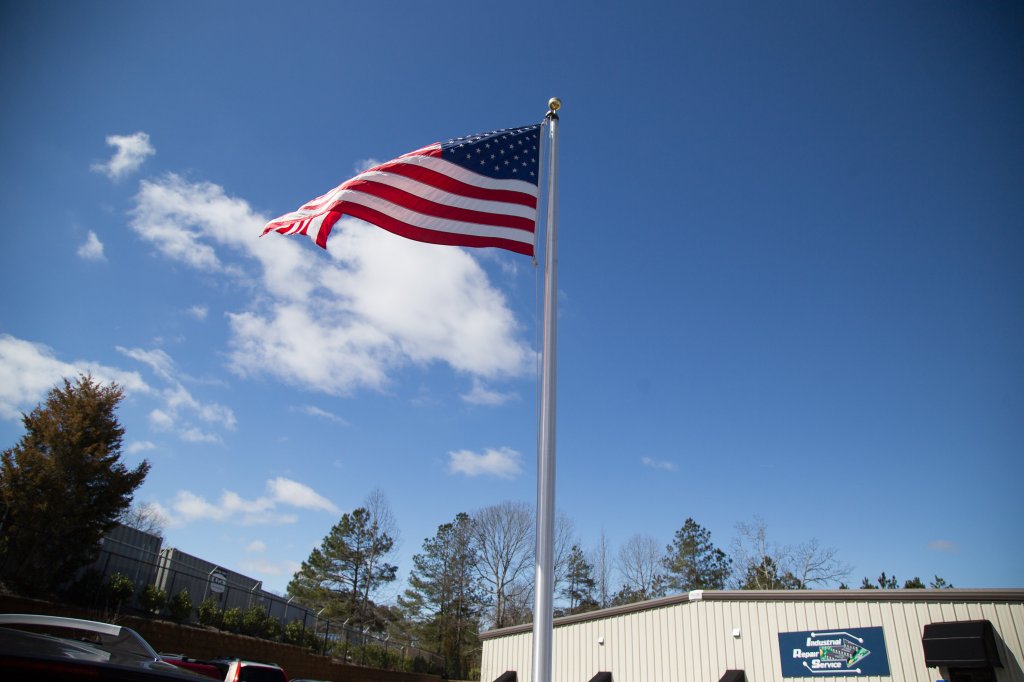 United States flag waving against a blue sky with scattered clouds, in front of a building marked 'Industrial Repair Service'.