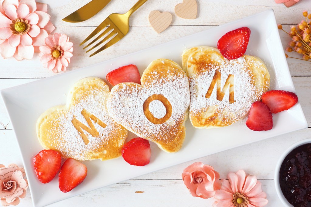 A plate of heart-shaped pancakes spelling 'MOM' topped with powdered sugar and accompanied by fresh strawberries and decorative flowers.