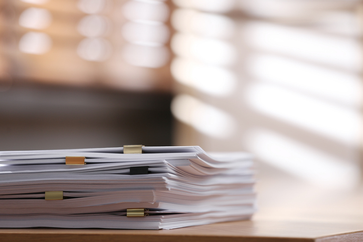 A stack of organized paperwork secured with binder clips, placed on a wooden surface, with soft light filtering through blinds in the background.