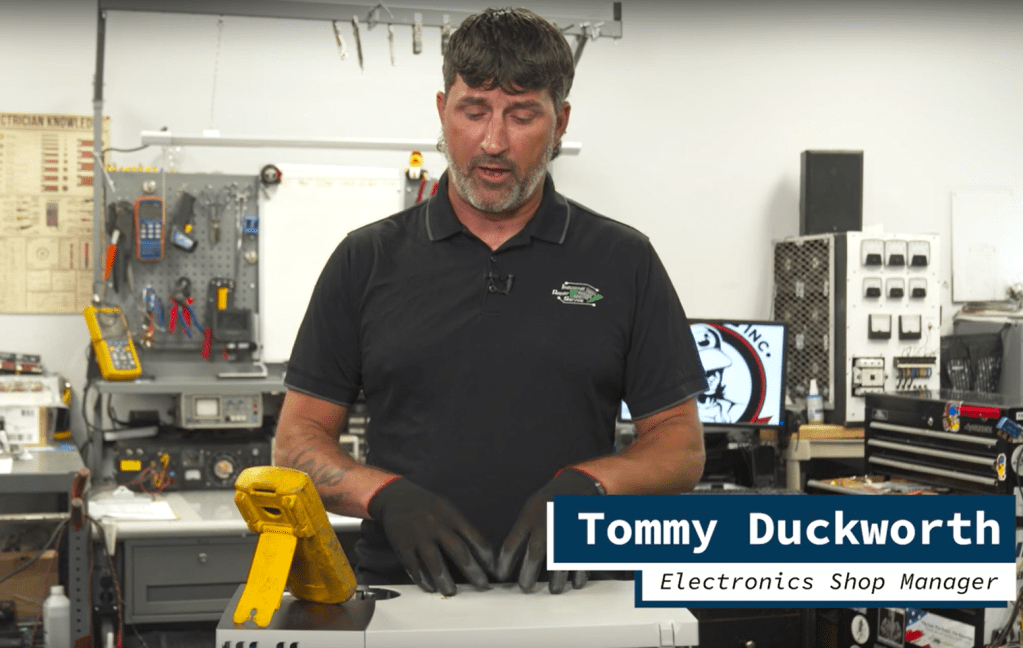 A man demonstrating a variable frequency drive (VFD) in a workshop, wearing gloves and standing in front of equipment and tools.