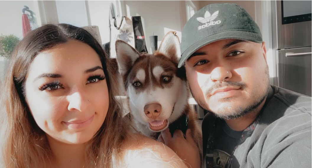 A smiling woman and man pose together for a selfie with a light brown and white husky dog, indoors with natural light.