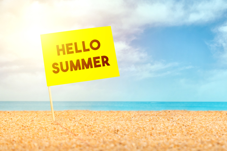 A bright yellow sign that reads 'HELLO SUMMER' stands on a sandy beach with a blue sky and ocean in the background.