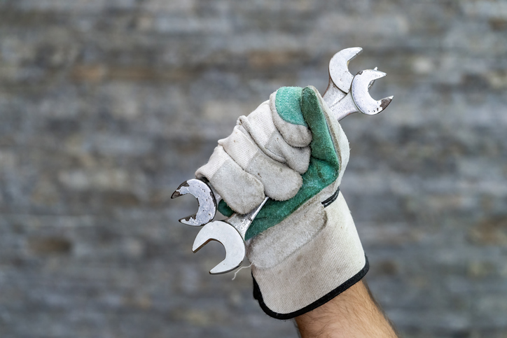 A gloved hand holding several metal wrenches, set against a blurred stone background.