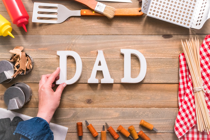 A hand placing white letters spelling 'DAD' on a wooden table, surrounded by barbecue utensils, condiments, and cooking accessories.