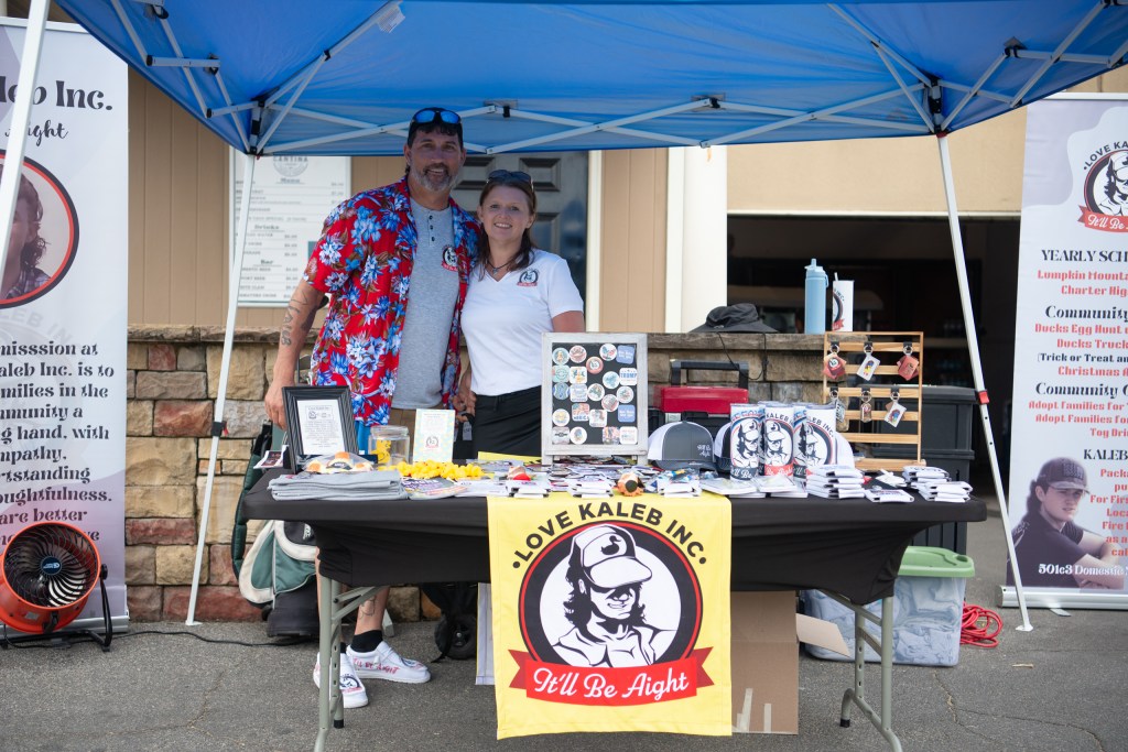 A fundraising booth for Love Kaleb Inc. featuring a man and a woman posing together with promotional materials, including merchandise and banners, under a blue tent.