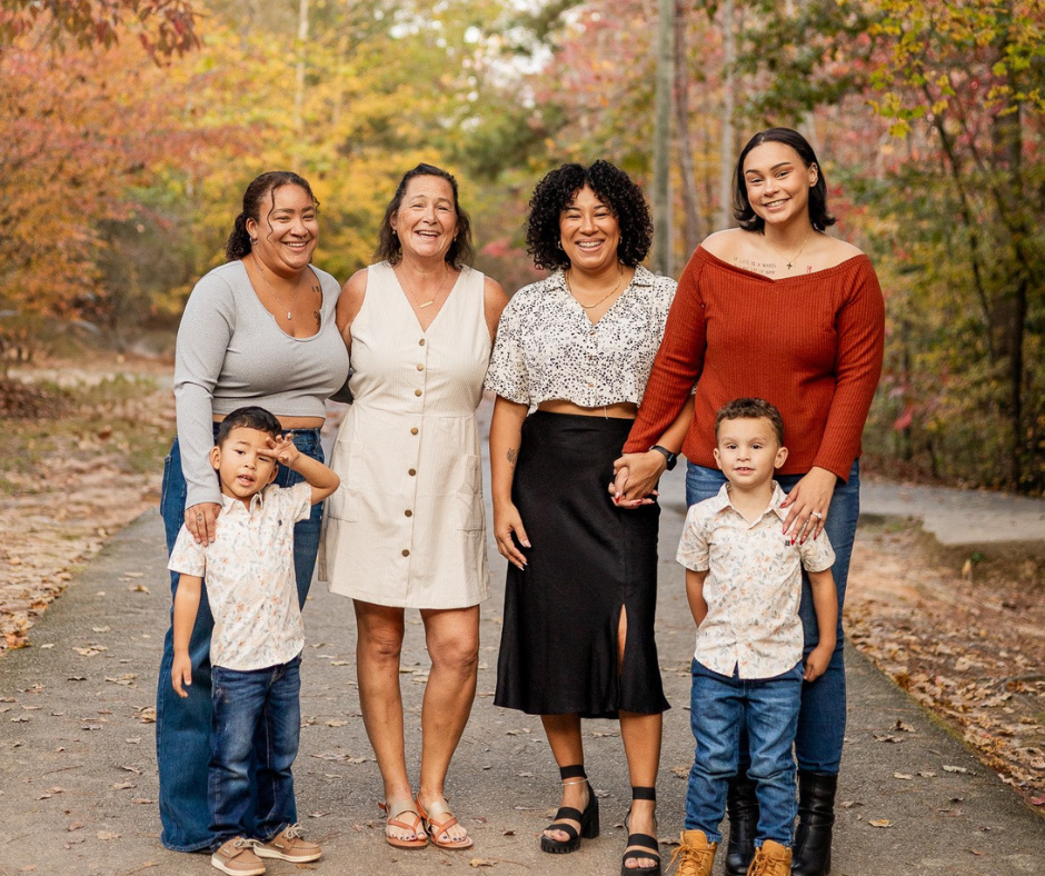 A group of five women and two young boys are standing together on a pathway surrounded by autumn foliage, smiling at the camera.