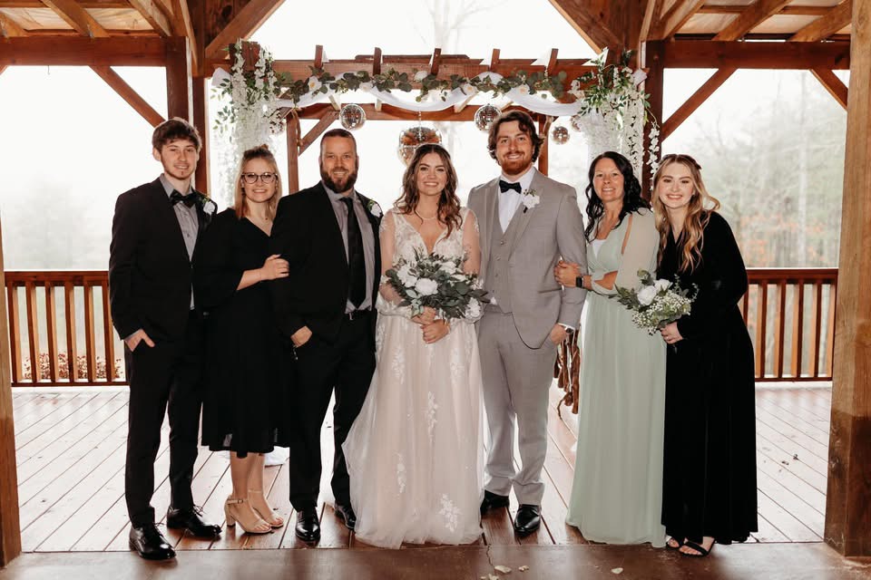 A group of six people posed together at a decorated venue, with a wooden structure and greenery in the background. They are dressed in formal attire, with the bride wearing a lace wedding gown and holding a bouquet, while others are dressed in elegant dresses and suits.