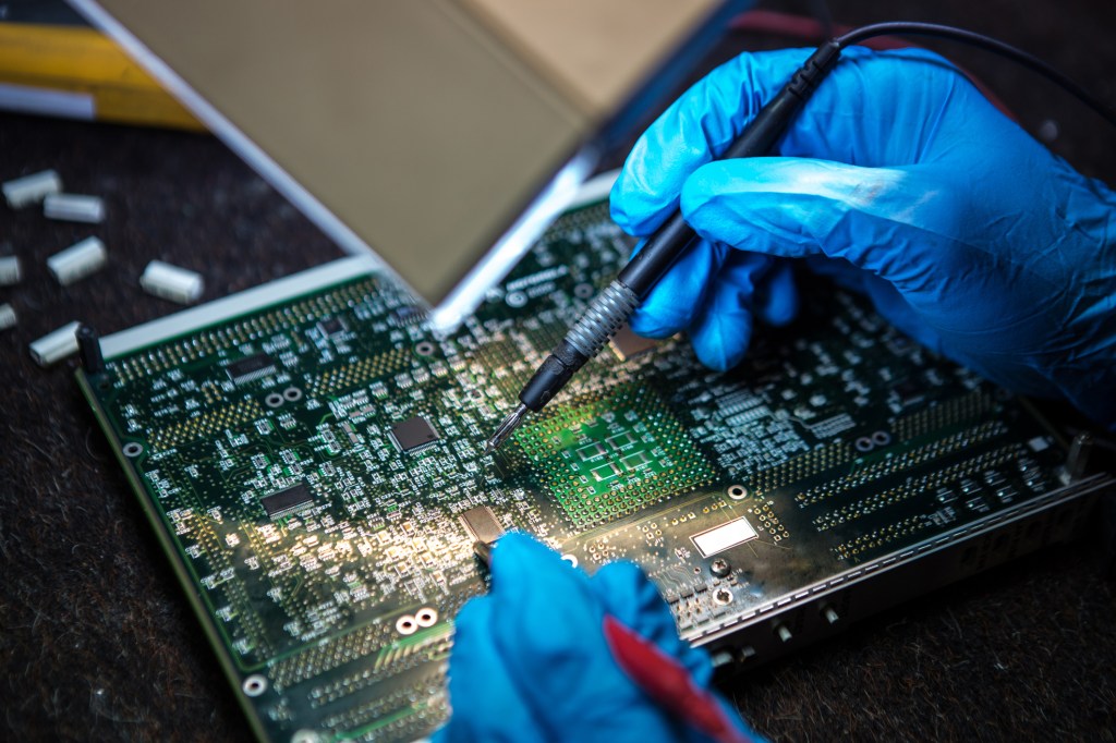 A technician wearing blue gloves repairing a printed circuit board (PCB) with a soldering tool, surrounded by small electronic components.