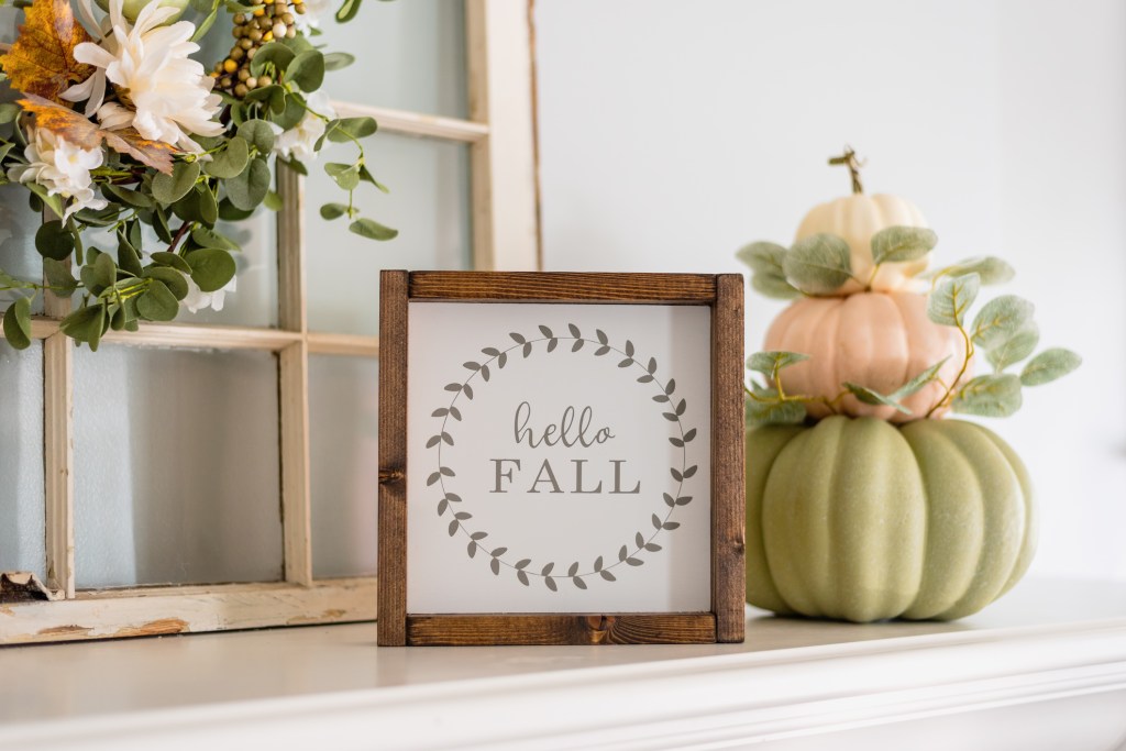 A decorative wooden frame displaying the text 'hello FALL' surrounded by a leaf design, next to a stack of pastel-colored pumpkins and greenery.