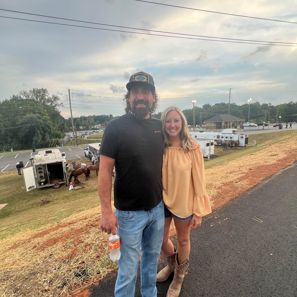 A man and a woman posing together outdoors, smiling. The woman is wearing a light-colored off-shoulder blouse and cowboy boots, while the man is in a black shirt and a cap. The background features vehicles and green areas, suggesting a casual outdoor setting.