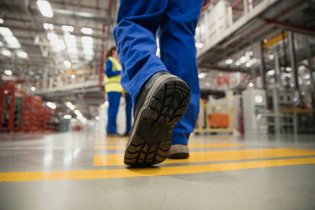 A worker in blue overalls walks through an industrial workspace, showcasing the sole of their safety boots on a yellow line.