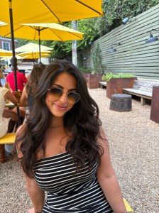 A woman with long dark hair wearing sunglasses, posing for a photo in a casual outdoor setting with yellow umbrellas and wooden furniture.