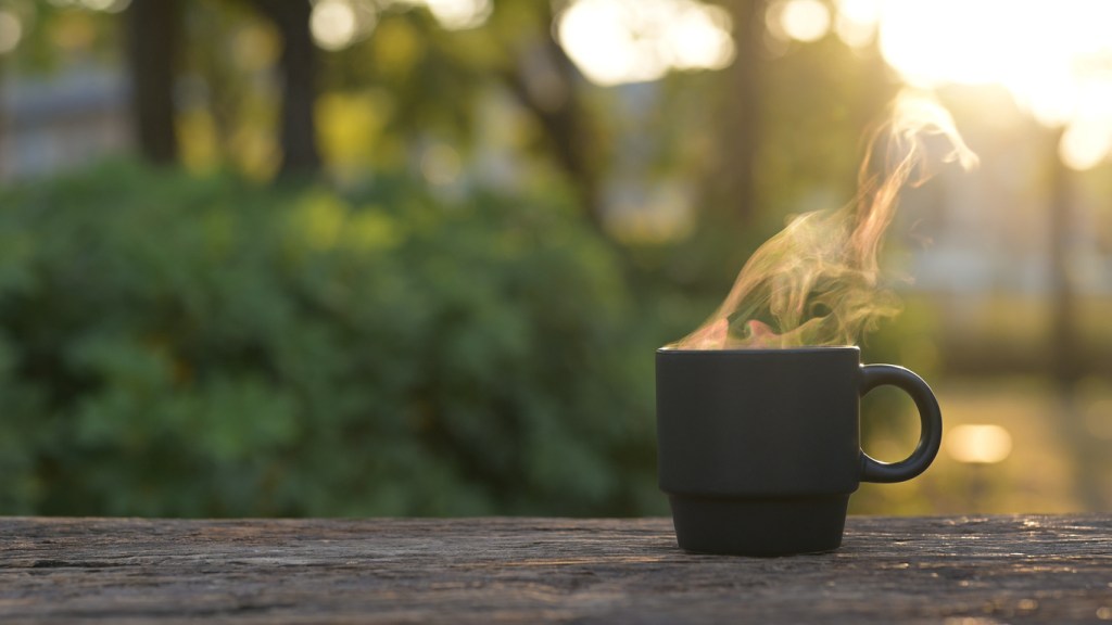A steaming black coffee mug placed on a wooden table with a blurred green background in the morning light.