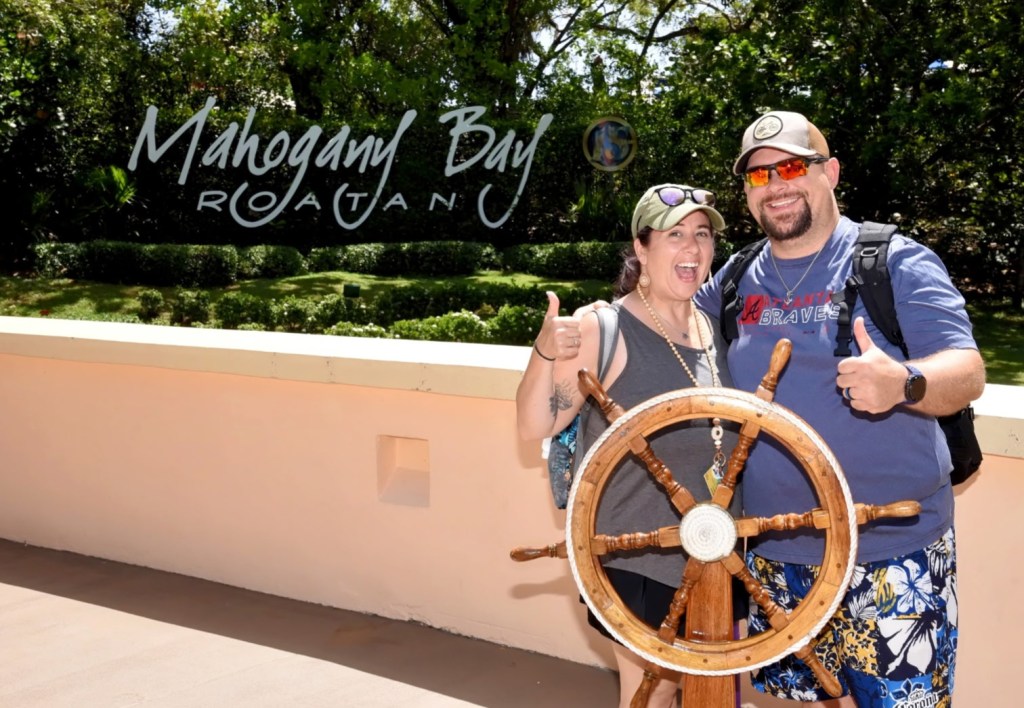 A joyful couple posing with a ship's wheel at Mahogany Bay, Roatan, both giving thumbs up.